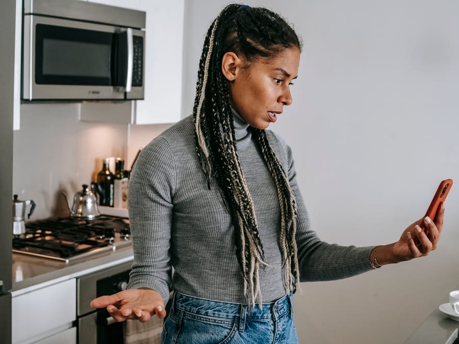 Serious young black lady with Afro braids in casual clothes gesticulating while having unpleasant conversation via video chat on smartphone in modern kitchen