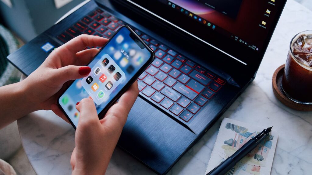 Hands holding a smartphone over a laptop with iced drink. Modern technology in everyday life.