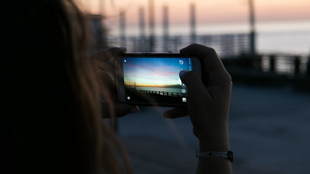 A person photographs a vibrant sunset with a smartphone by the waterfront. Outdoor scene.