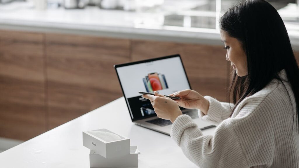 Young Asian woman setting up a new smartphone while working on a laptop in a modern indoor setting.