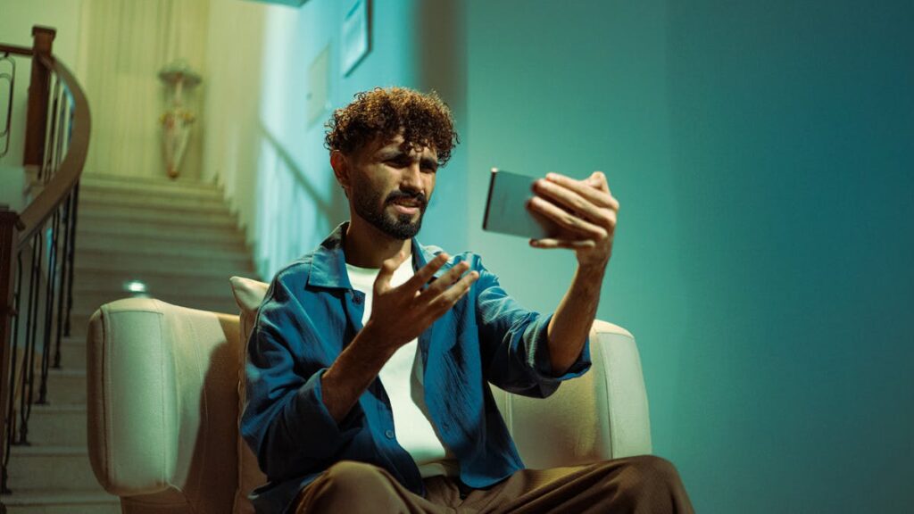 A man sitting on a couch indoors, looking confused while using a smartphone.