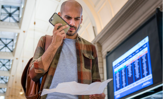 Man talking on a smartphone while reading a document near a digital travel information board