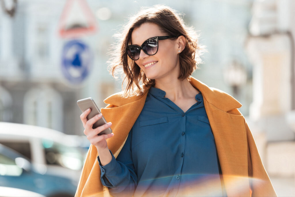 Smiling woman wearing sunglasses and holding a smartphone while walking outdoors in the city.