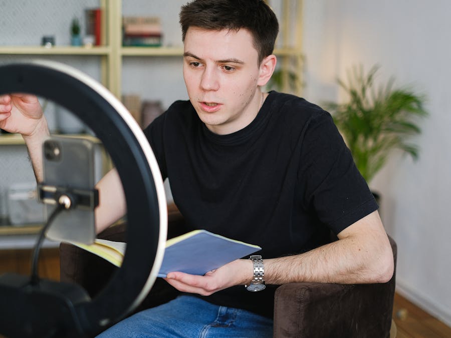 Adult male recording video content indoors, using a smartphone and ring light, holding a notebook.