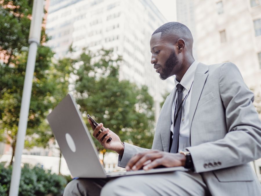 Businessman in a suit using a laptop and smartphone in an urban setting, blending technology with business.