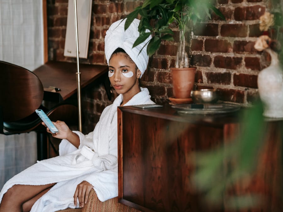 Side view of young female in bathrobe and towel on head browsing smartphone while sitting on chair at home