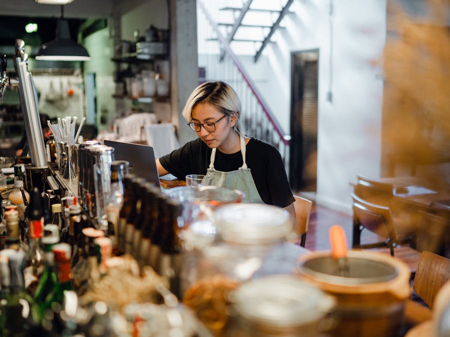 Focused young ethnic woman in apron and eyeglasses sitting at bar counter full of various drinks and using laptop