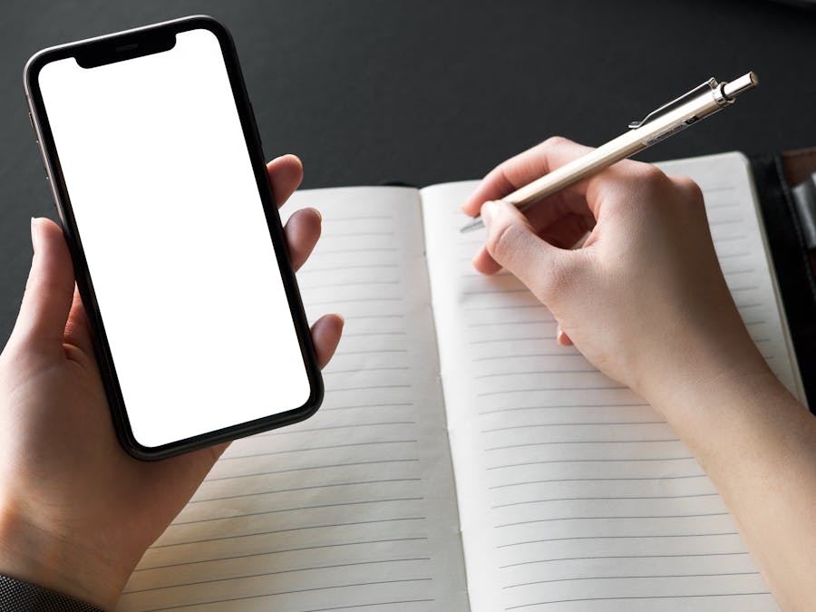 Person writing in a notebook while holding a smartphone with a blank screen, ideal for mockups.