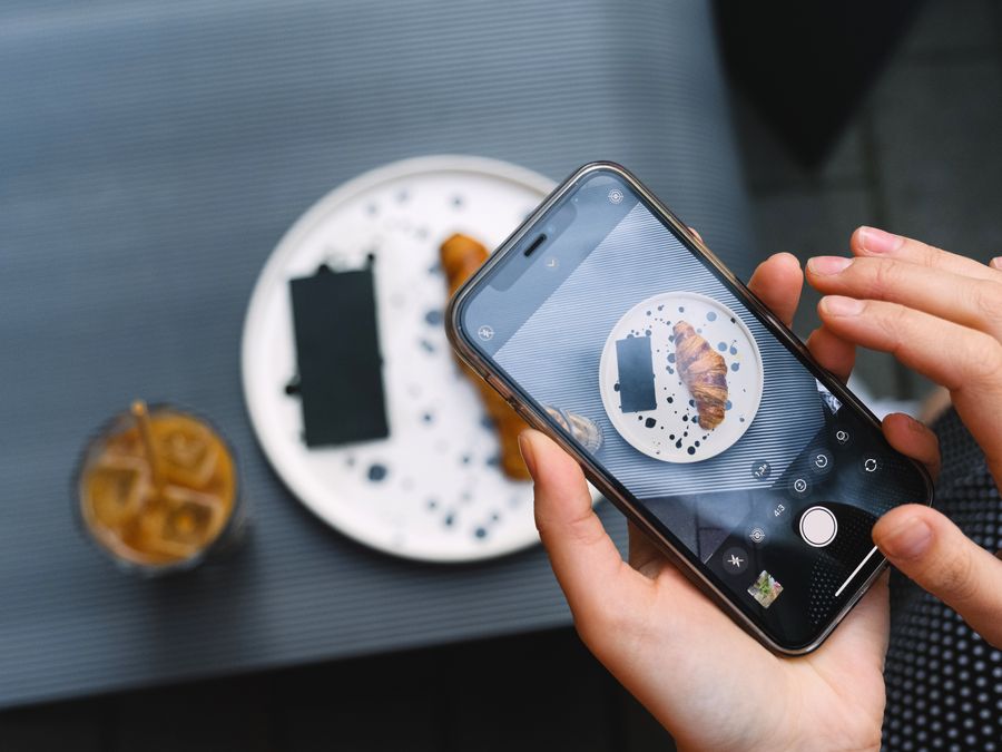 Hands photographing a croissant on a plate with a smartphone, featuring coffee.