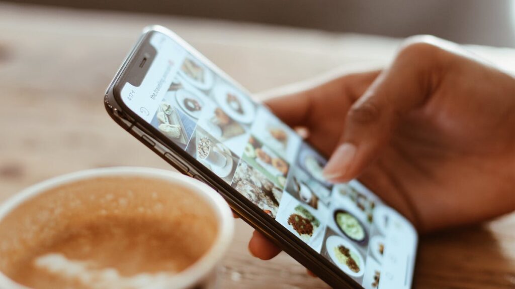 Hand browsing social media photos on a smartphone next to a cup of coffee indoors.