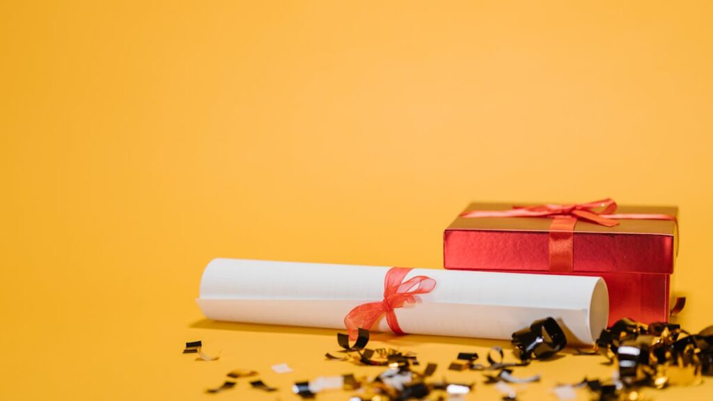 A red gift box and diploma with ribbons on a vibrant yellow background symbolizing celebration and achievement.