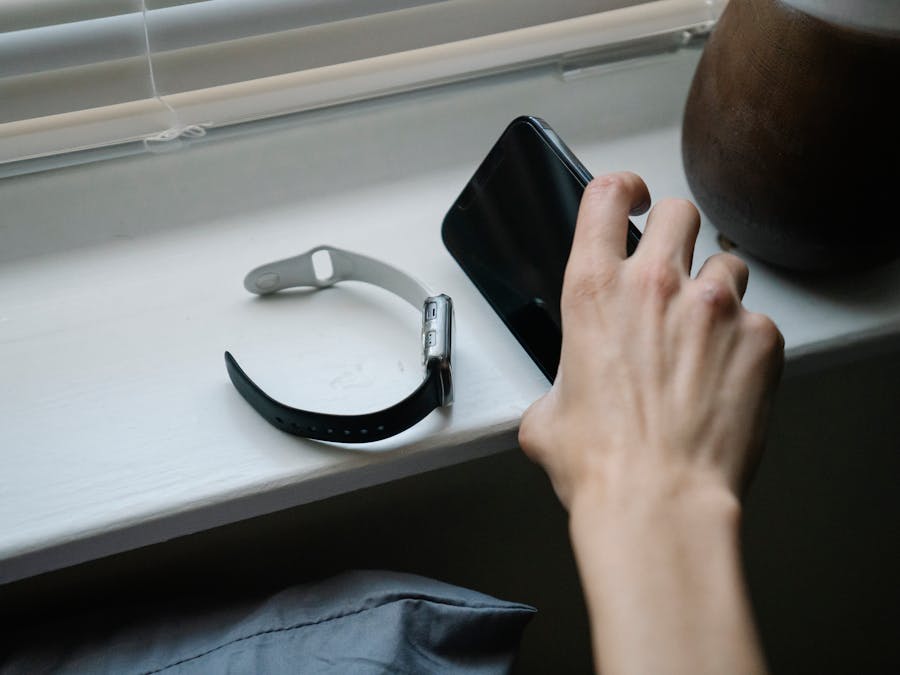 From above of crop anonymous person taking cellphone placed on windowsill near smart watch while waking up in morning