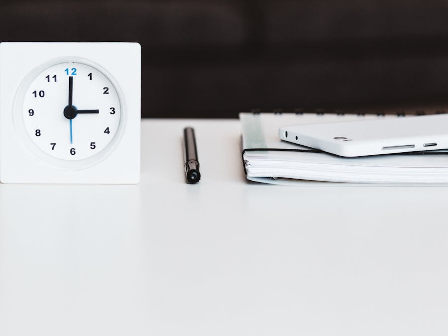 A minimalist office desk setup with a clock, pen, smartphone, and notebook, conveying time management and organization.