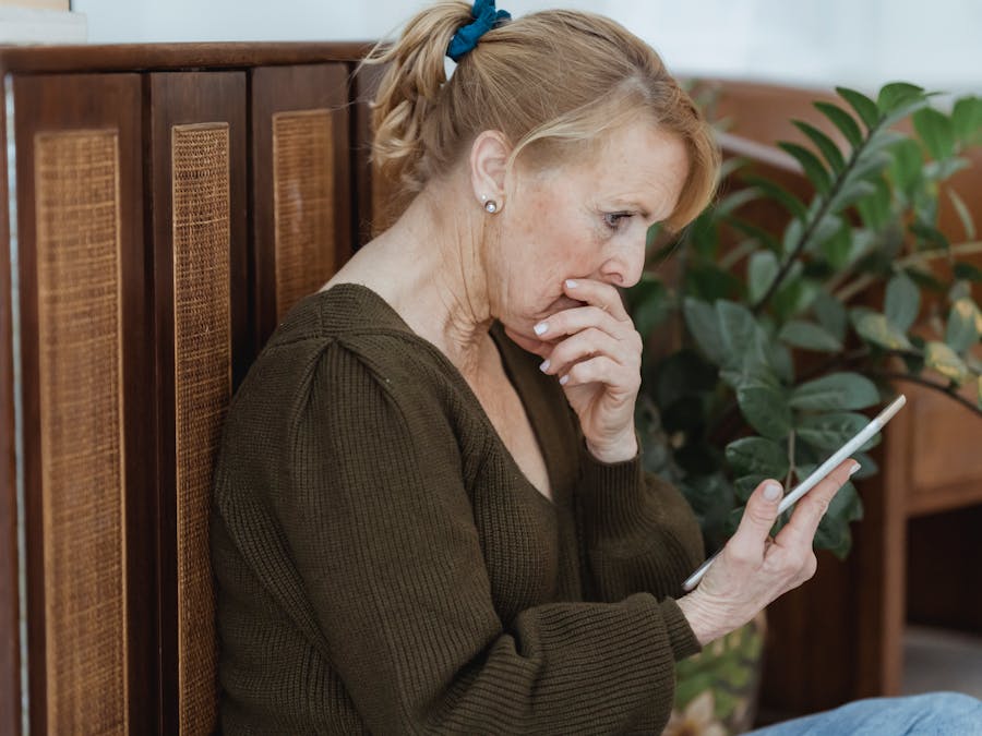 Side view of surprised elderly female with cellphone covering mouth against cabinet and plant at home