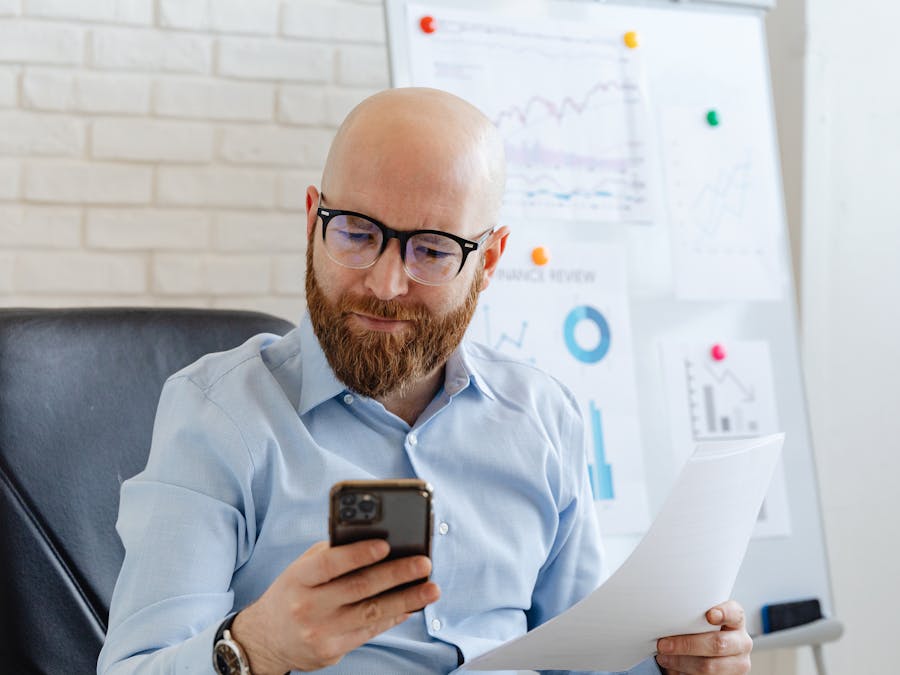 Bald businessman with eyeglasses checking phone data during an office presentation.