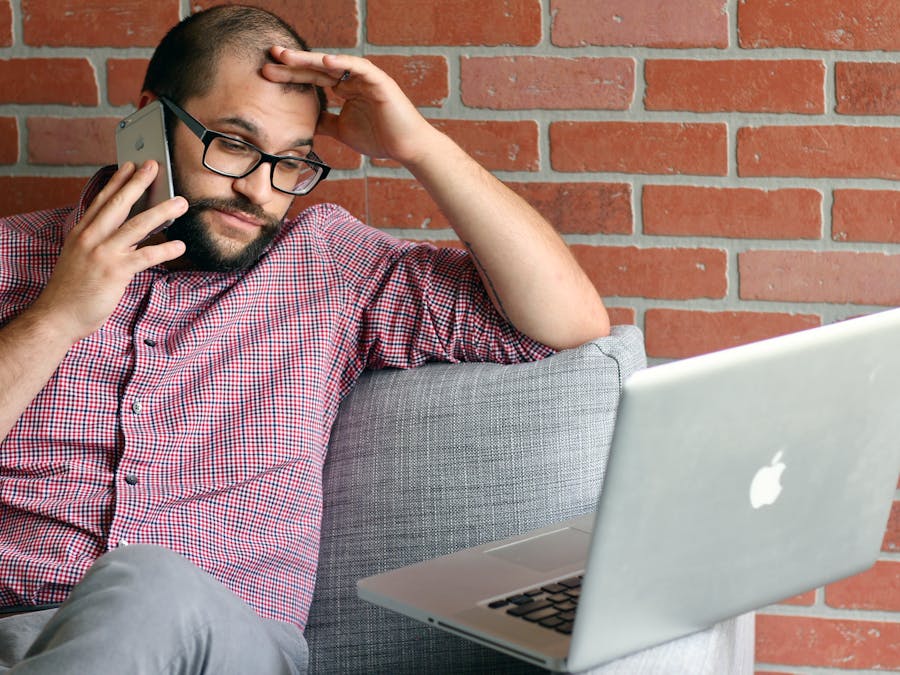 Bearded man looks frustrated while working remotely with laptop and phone.