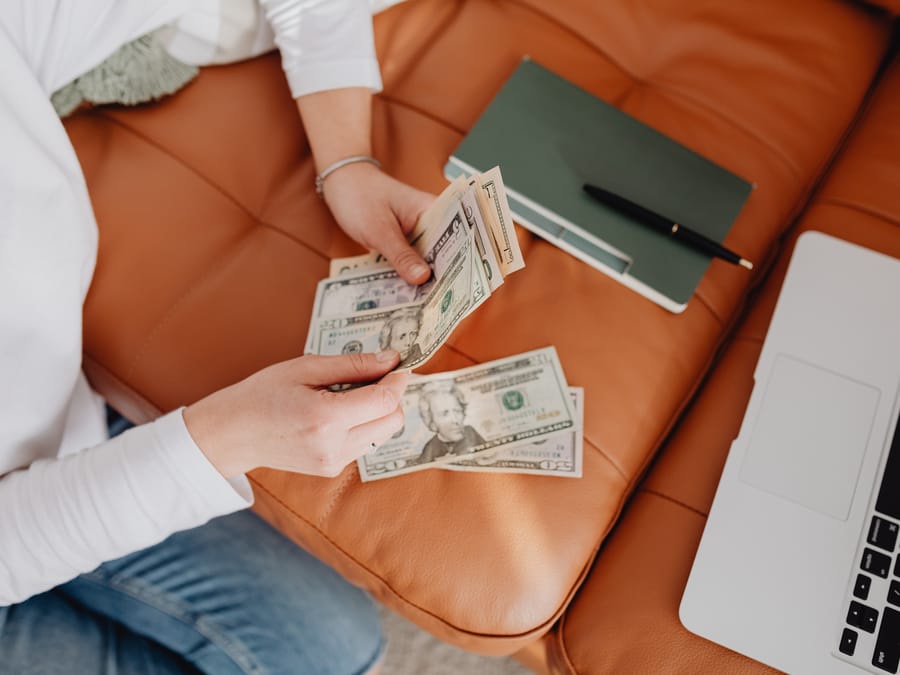 Person counting dollar bills on a sofa next to a notebook and laptop.