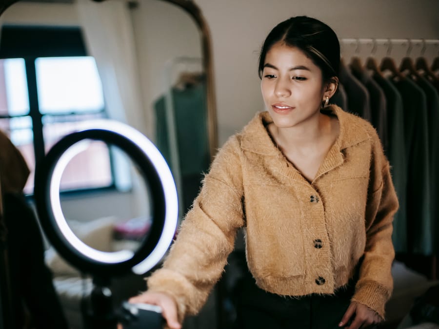 Confident young ethnic lady in trendy outfit standing in wardrobe with smartphone on ring light while taking selfie near mirror