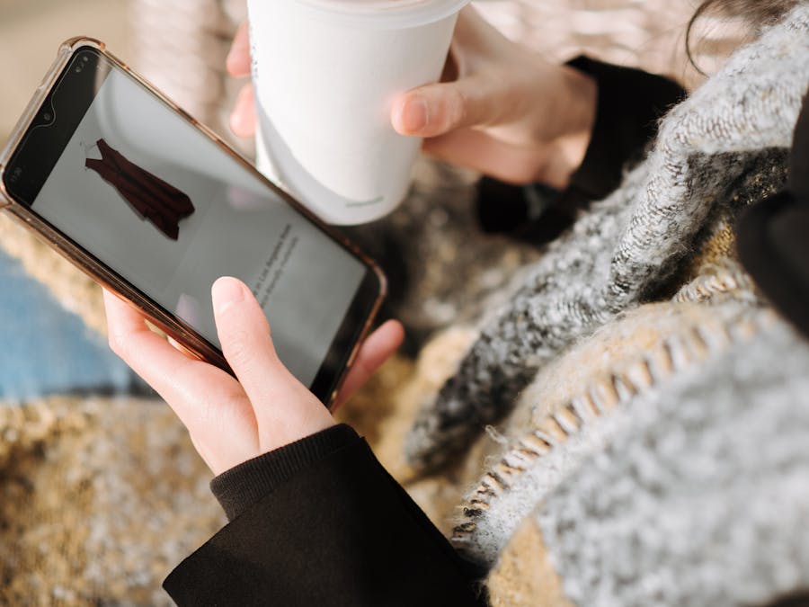 From above of crop anonymous female purchaser with takeaway hot drink choosing dress on cellphone screen during online shopping