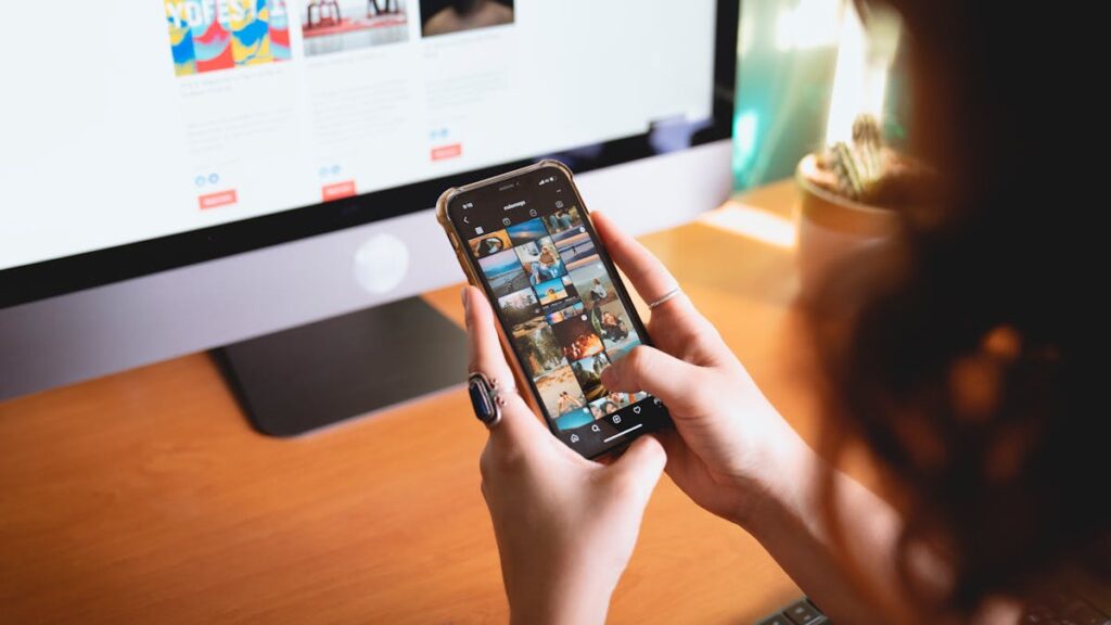 A person browsing images on a smartphone while sitting at a desk with a desktop monitor.