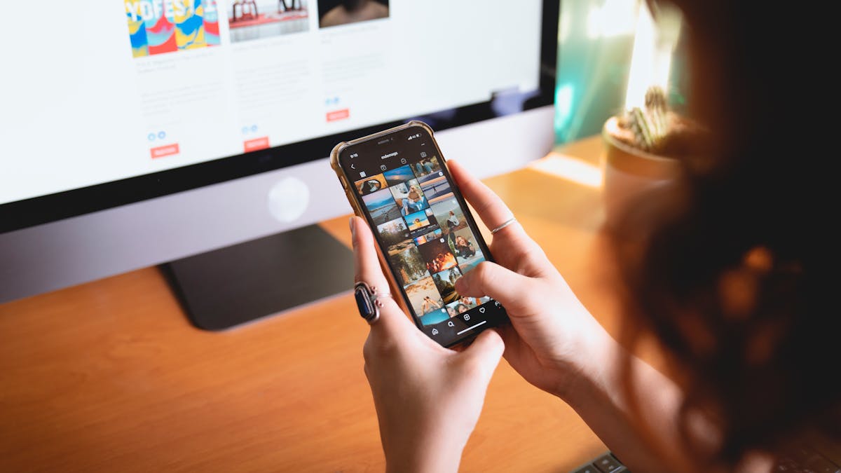 A person browsing images on a smartphone while sitting at a desk with a desktop monitor.
