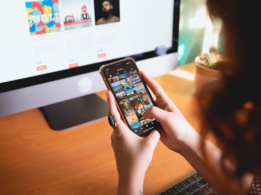 A person browsing images on a smartphone while sitting at a desk with a desktop monitor.