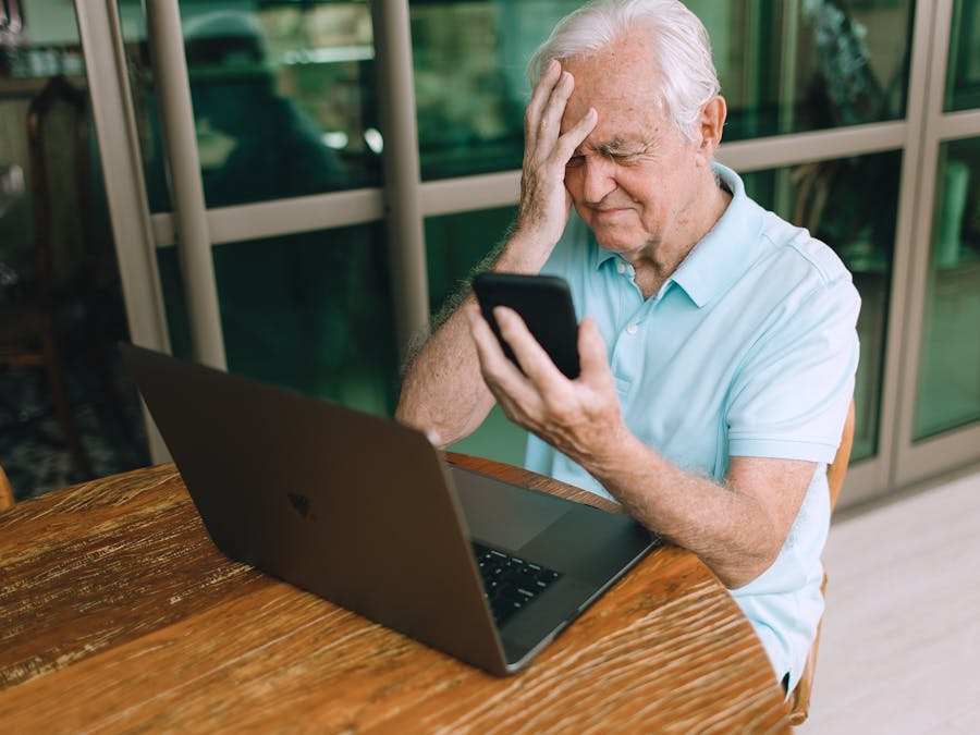 Elderly man frustrated by smartphone and laptop technology indoors at home.