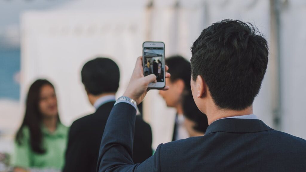 Man in suit photographing colleagues with smartphone during business event outdoors.