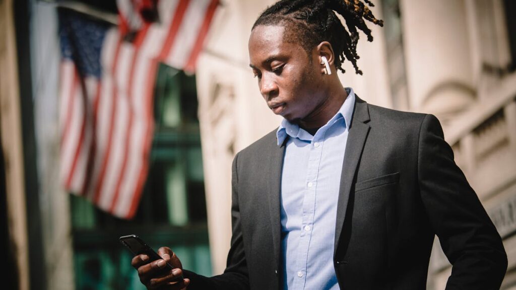 Low angle of young African American male entrepreneur in formal suit messaging on cellphone with wireless earbuds in ears while standing against blurred facade of building with American flags
