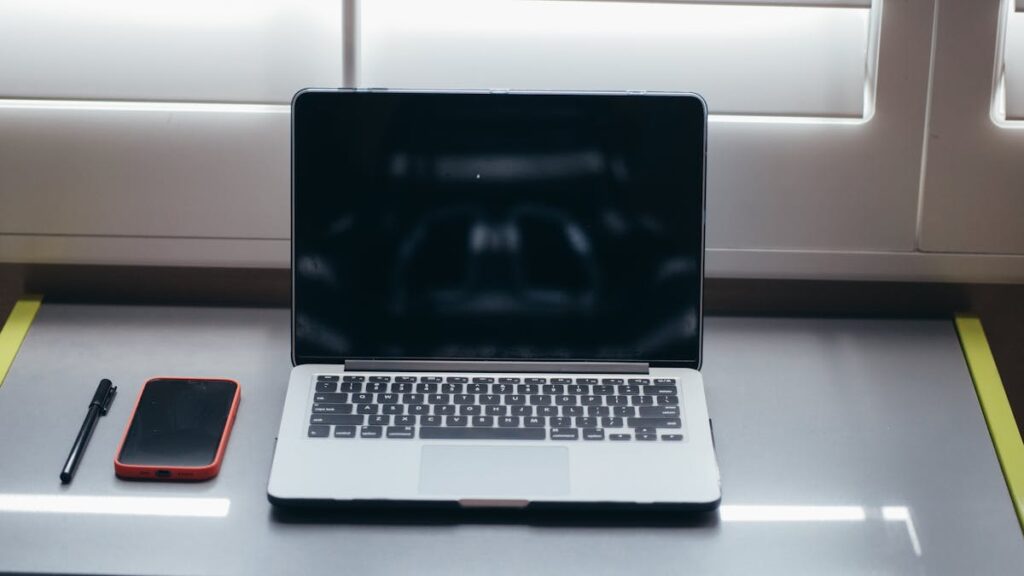 A minimalist desk setup featuring a laptop, smartphone, and pen under soft window light.