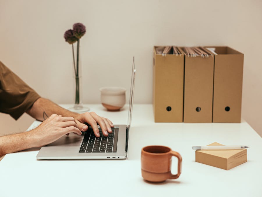 Hands typing on a laptop in a minimalist workspace with coffee and file organizers.