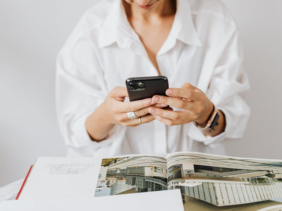 Woman in white shirt using smartphone while reading architecture magazine. Indoor setting.