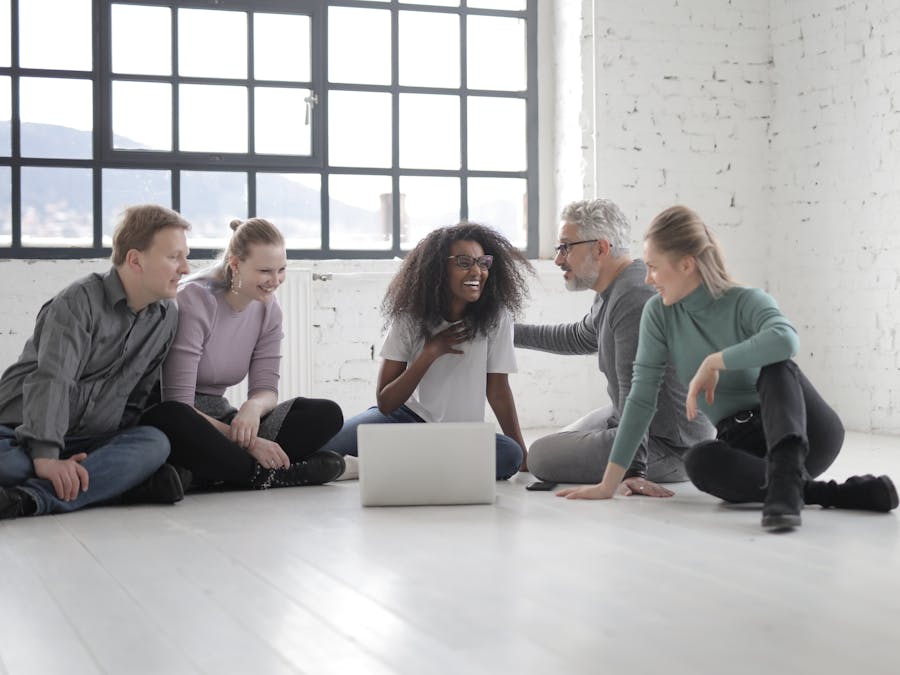 Happy multiethnic group of people gathering together while speaking and sitting with crossed legs in front of portable computer on ceramic floor near window in sunlight
