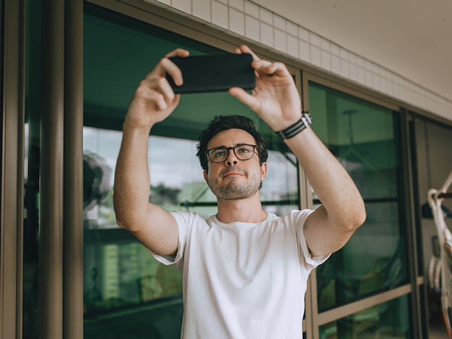 A young man with glasses takes a selfie on a modern balcony using a smartphone, looking upward.
