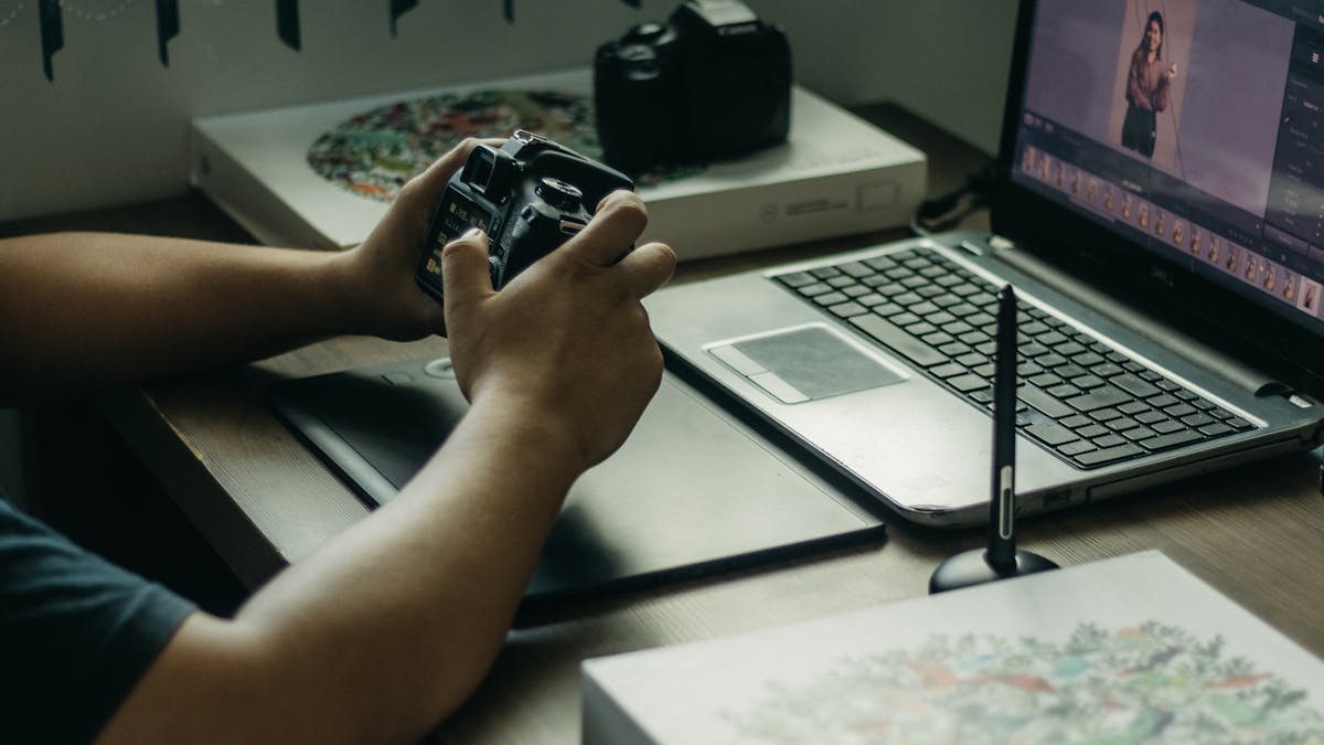 Photographer using a laptop and stylus for photo editing, holding a camera in an indoor setup.
