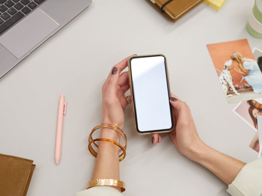 Close-up of a woman holding a smartphone on a desk with a laptop and photos, capturing modern workspace vibes.