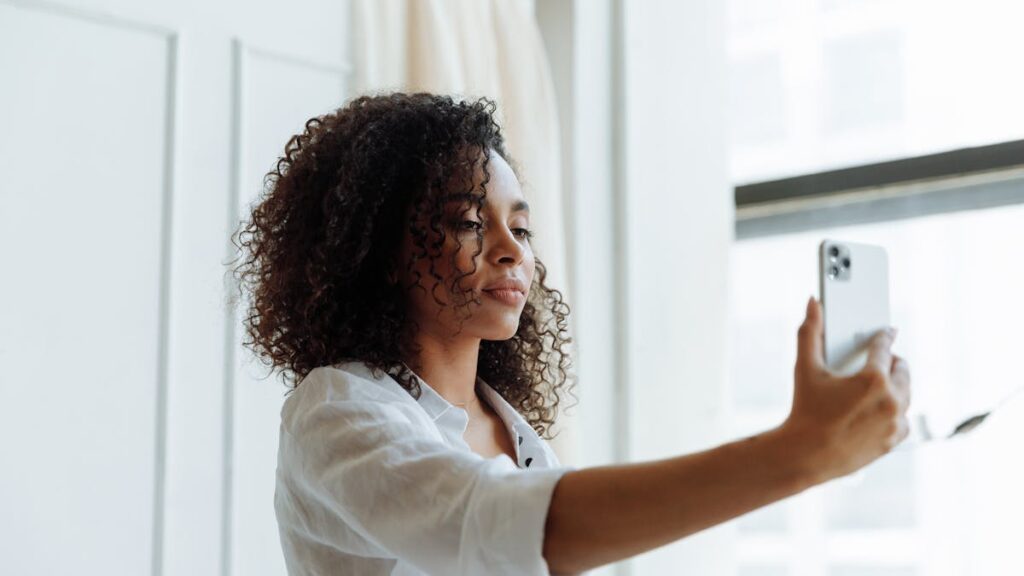 Young woman with curly hair taking a selfie with smartphone indoors.