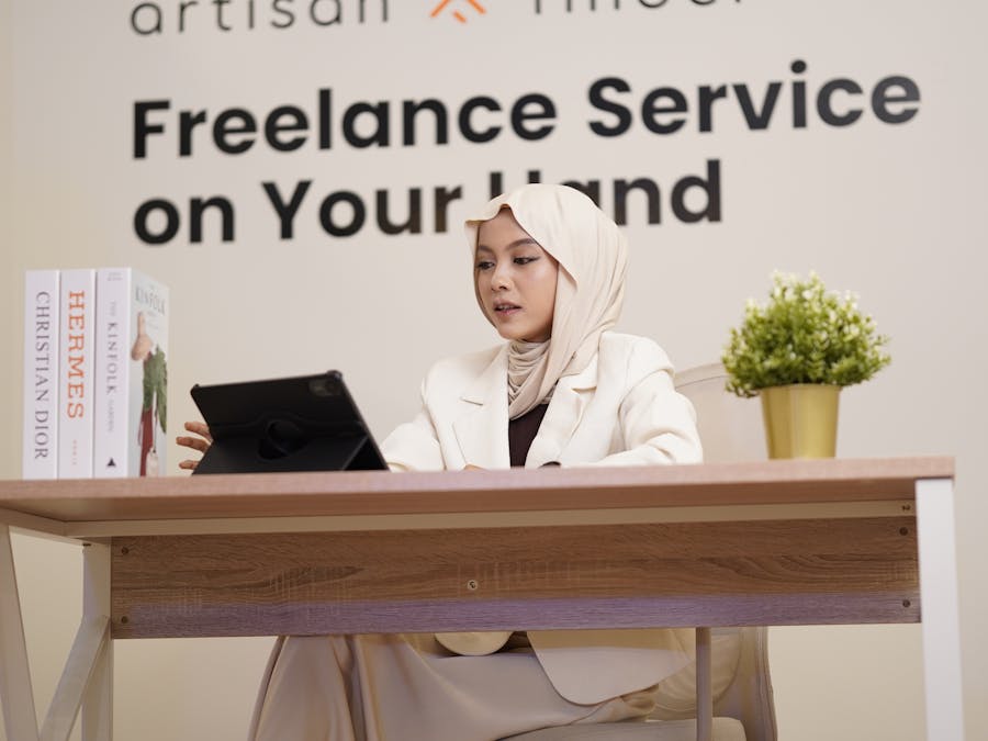 Professional woman in hijab working on a tablet at a modern office desk with books and decor.
