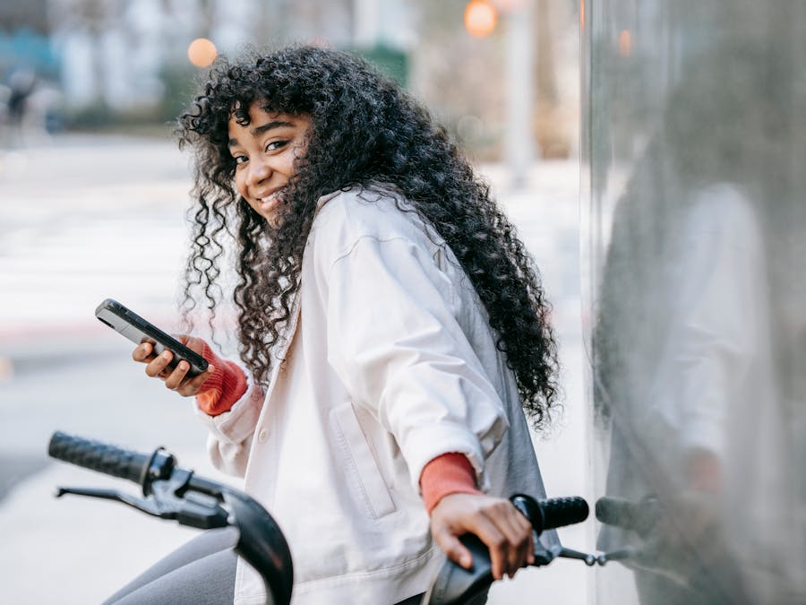 Side view happy African American female in casual clothes standing on sunny city street with bicycle and using modern mobile phone while looking at camera with toothy smile