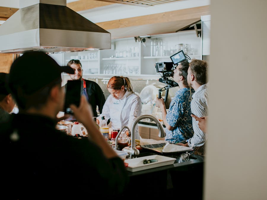 Behind the scenes view of film crew capturing a cooking show in a professional kitchen setting.