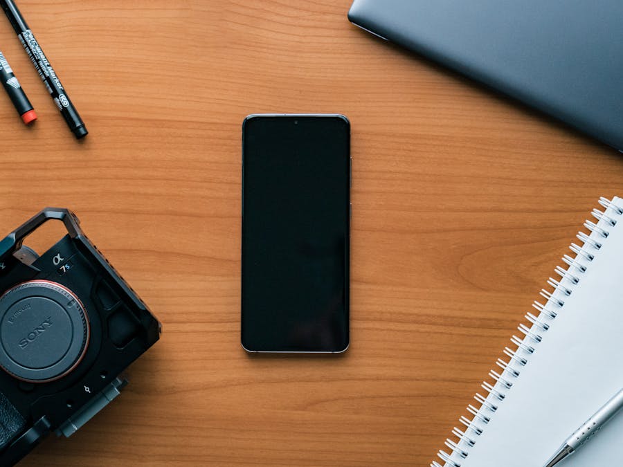 A neat flat lay of a modern workspace featuring a smartphone, camera, pens, and notebook.