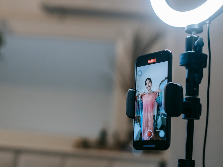 Anonymous female blogger standing in room and shooting vlog on smartphone with ring lamp while demonstrating pink dress in hands