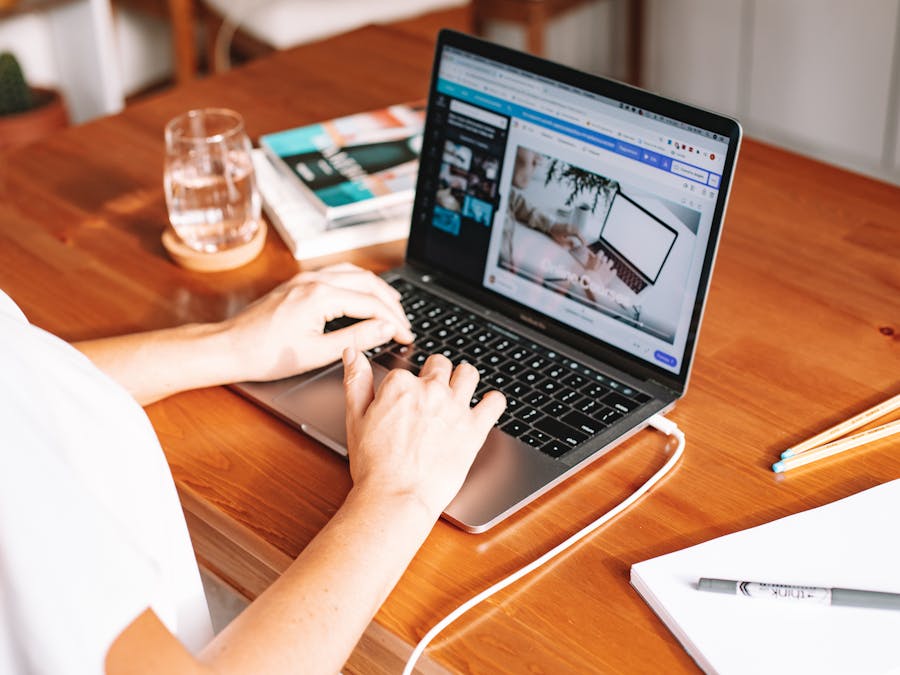 A person working remotely on a laptop at a wooden desk with stationery and a notepad.