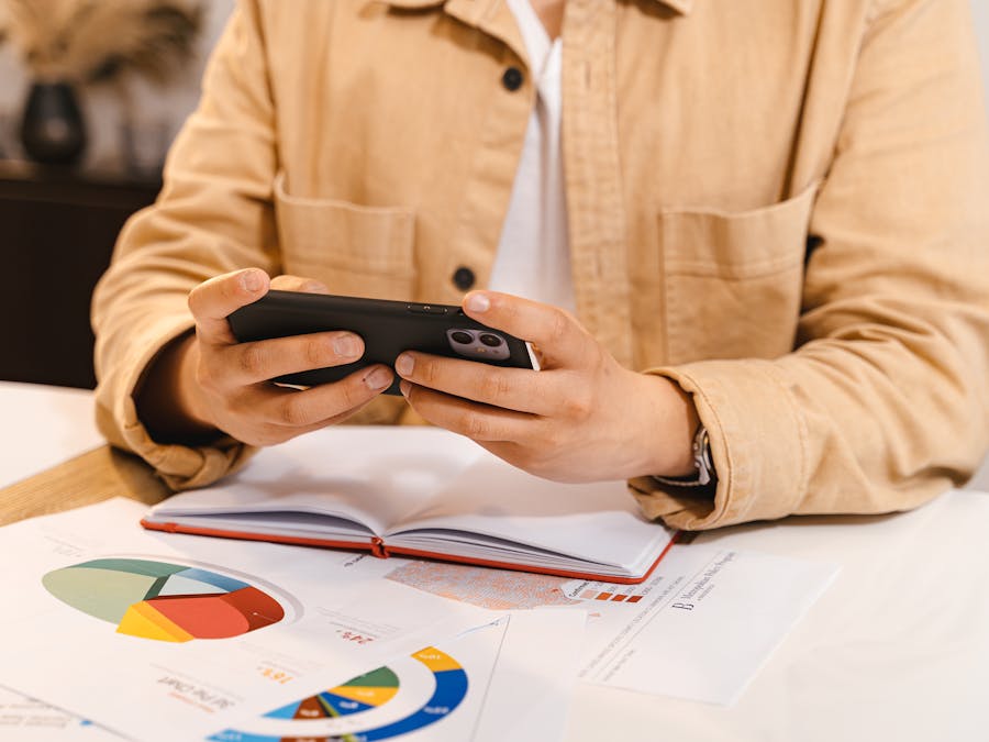 Person in jacket using smartphone, reviewing charts and data at a workspace.