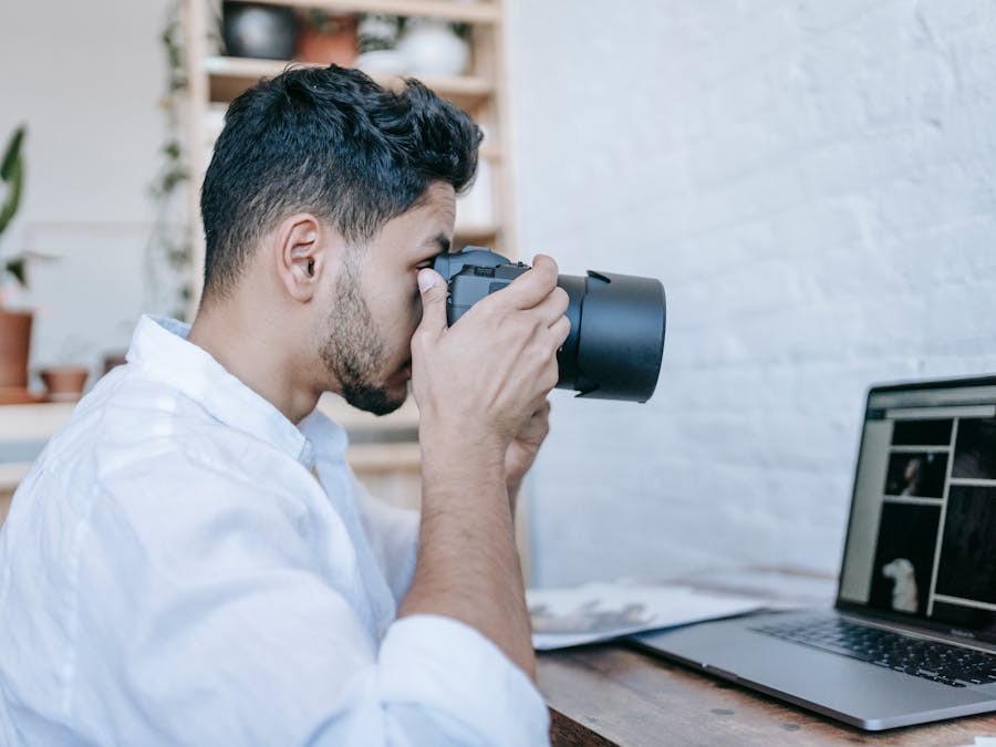 Side view of young bearded male photographer taking picture on professional photo camera while sitting at table with laptop in modern loft style studio