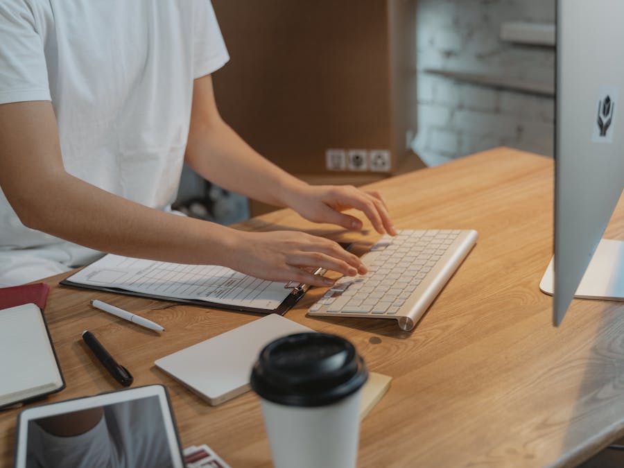A person typing on a keyboard with documents on a wooden desk. Modern workspace setup.