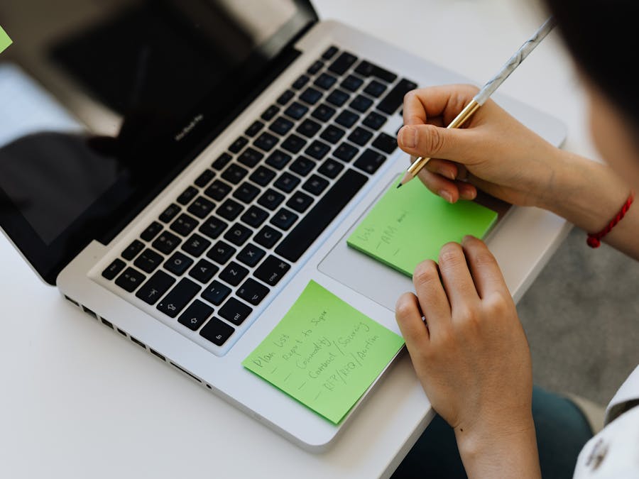 An individual writes notes on green sticky notes placed on a laptop in a work setting.
