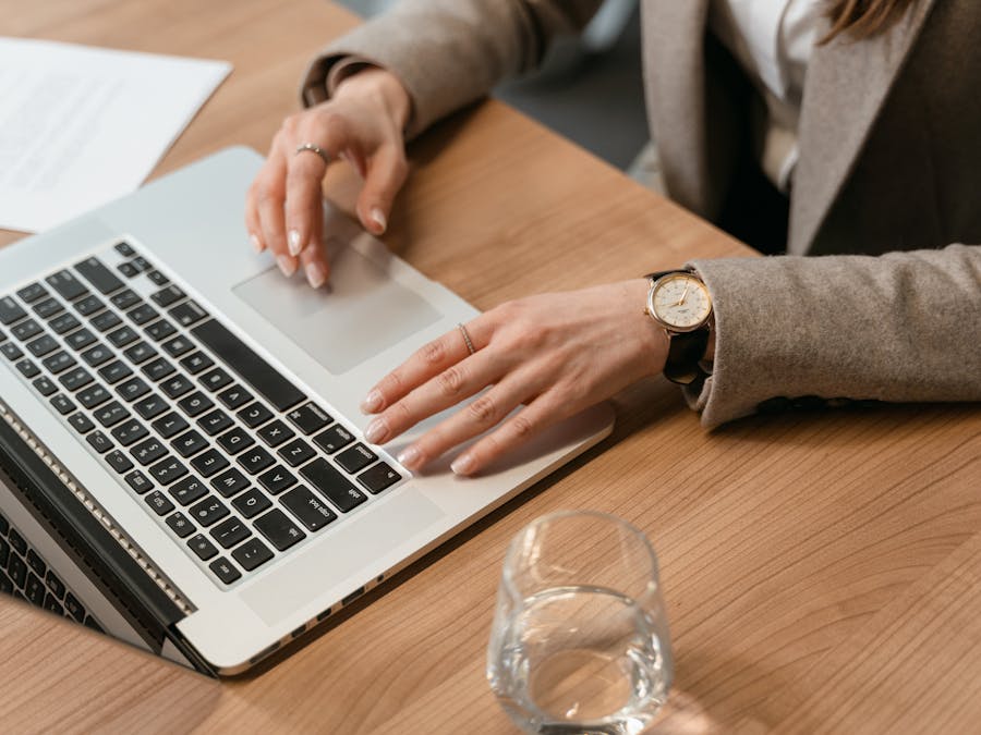 Woman working on a laptop at a wooden desk with a glass of water and documents.
