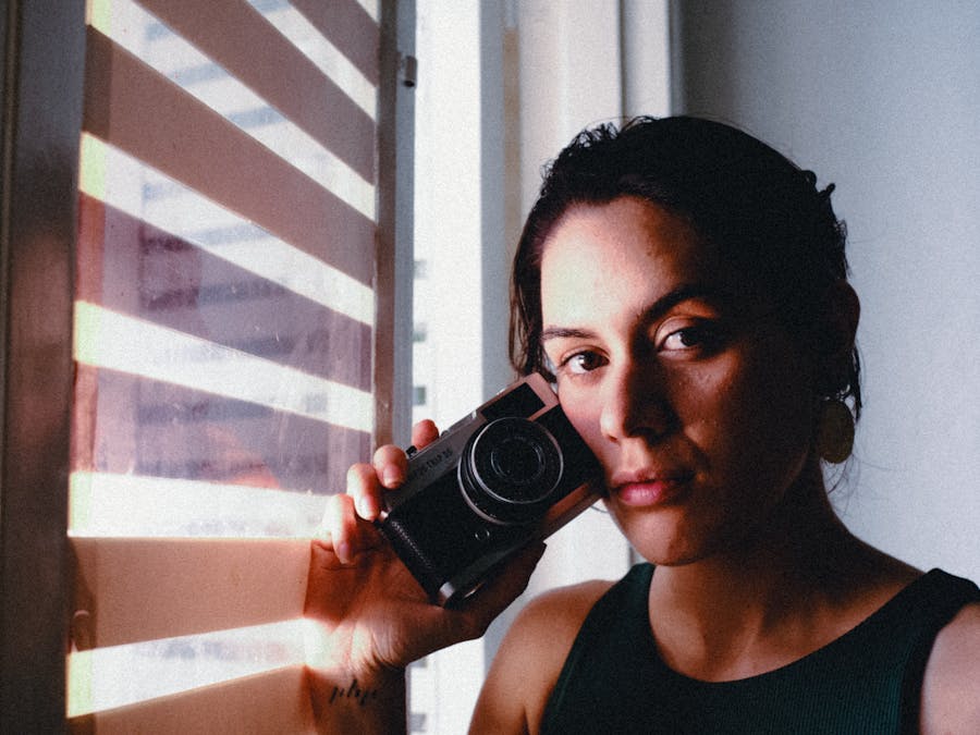 Portrait of a woman holding a camera by a sunlit window, showcasing artistic lighting and moody ambiance.