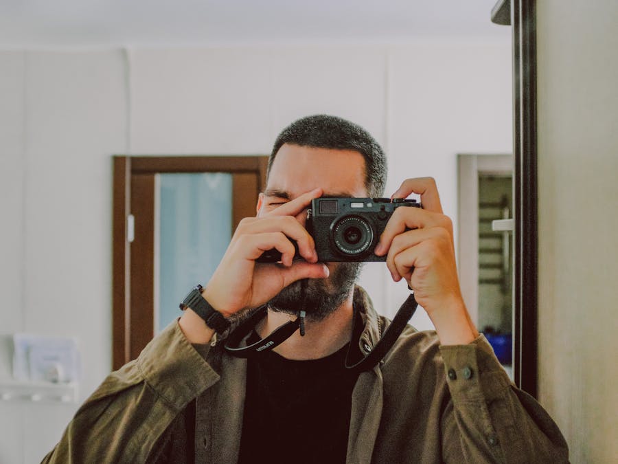 Bearded guy using photo camera to take selfie near mirror in cozy room at home